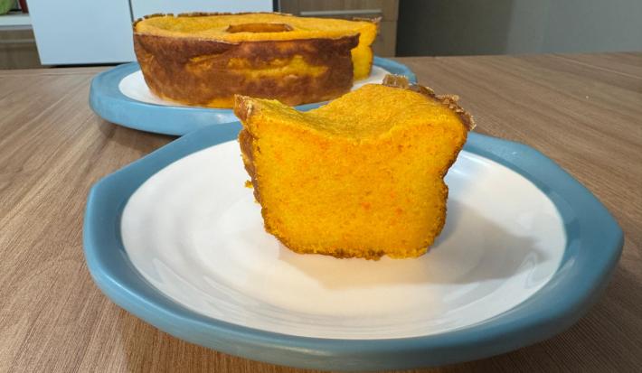 Fotografia em tons claros com um bolo de cenoura mole ao centro. O bolo de cenoura tem cor laranja, textura lisa e cremosa