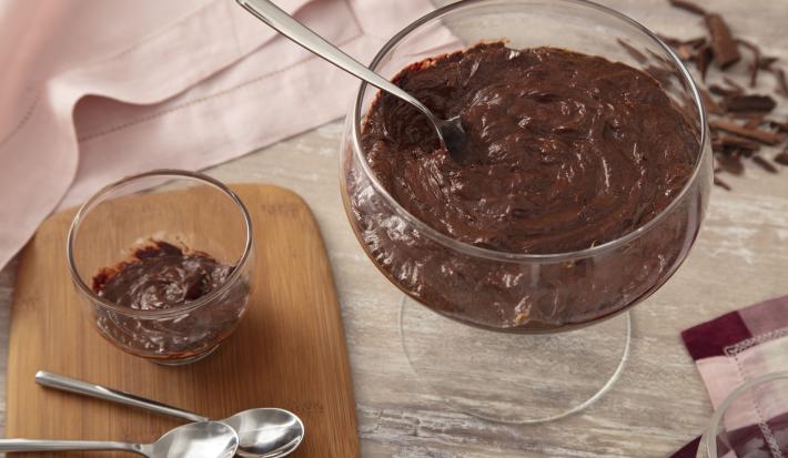 Foto da receita de Mousse Fácil de Chocolate. Observa-se um recipiente redondo com a mousse e, ao lado, um recipiente individual. Também tem duas colherinhas ao lado e raspas de chocolate como decoração.