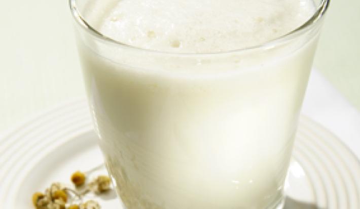 Fotografia em tons de branco e bege em uma bancada de madeira branca, um prato branco, um copo de vidro alto com a bebida com Leite Molico e chá de camomila.