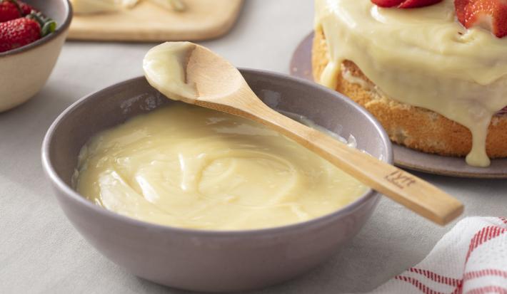 Fotografia em tons de vermelho em uma bancada de madeira cinza, um pano branco com listras vermelhas, um recipiente cinza com a ganache de chocolate branco. Ao fundo, um bolo coberto com a ganache e decorado com morangos. Fotografia em tons de vermelho em uma bancada de madeira cinza, um pano branco com listras vermelhas, um recipiente cinza com a ganache de chocolate branco. Ao fundo, um bolo coberto com a ganache e decorado com morangos.