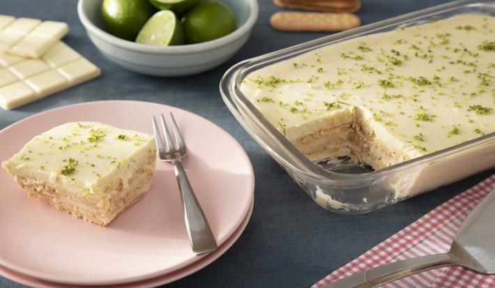 Foto da receita de Pavê de Leite Moça e Limão com Chocolate Branco. Observa-se uma fatia do pavê em um prato de sobremesa rosa ao lado da travessa inteira de vidro com a sobremesa. Atrás, um recipiente com limões cortados e biscoito Tostines decoram a fot