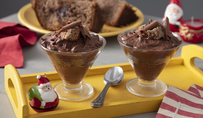 Fotografia em tons de amarelo e vermelho em uma bancada de madeira com decoração natalina, ao centro uma bandeja amarela com duas taças recheadas com chocolate e panettone e um paninho vermelho listrado ao lado.