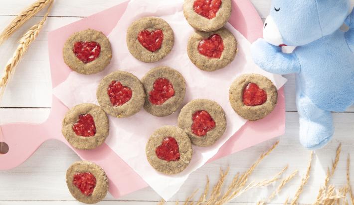 Foto da receita de Biscoitos de Coração. Observa-se uma tábua rosa bebê com os biscoitos dispostos sobe um papel manteiga. Alguns trigos e o ursinho de Mucilon decoram a foto.