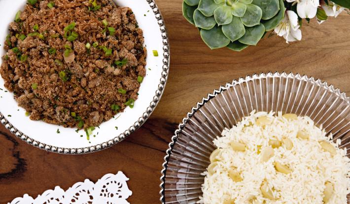 fotografia em tons de marrom e branco tirada de uma bancada marrom vista de cima. Contém um recipiente ao lado esquerdo redondo branco com a farofa e ao lado direito um recipiente redondo transparente com arroz