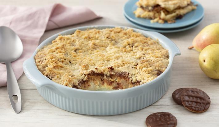 Fotografia em tons de azul em uma bancada de madeira clara, um paninho bege, um recipiente azul claro com o crumble de pera com calipso. Ao fundo, um prato com um pedaço do crumble, peras e calipso espalhados pela mesa.