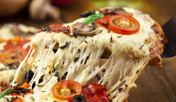 Fotografia de uma pizza com o queijo puxando e bem recheada, como queijo, azeitona, tomates e ricota. A pizza está sobre uma tábua de madeira de toma escuro, e ao fundo, cebola, tomate e champignon.