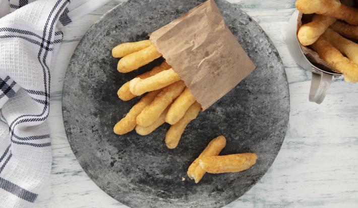 Fotografia em tons de azul em uma bancada de madeira azul mesclada, um pano branco com listras azuis, um prato redondo cinza com vários biscoitos de polvilho fritos. Ao lado, uma caneca com mais biscoitos dentro.