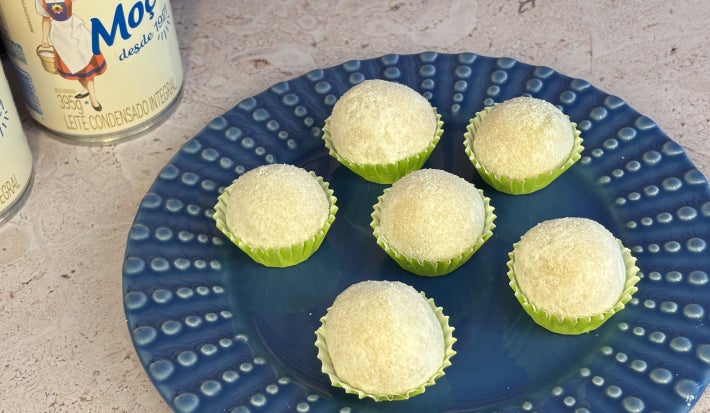 Seis brigadeiros brancos em forminhas verdes sobre prato azul decorado, com latas de LEITE MOÇA® ao fundo.