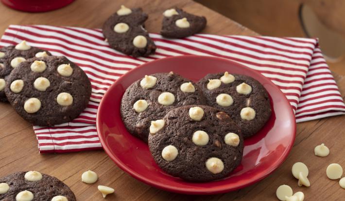 Fotografia em tons de vermelho em uma bancada de madeira com um pano branco com listras vermelhas, um prato vermelho ao centro com três cookies de chocolate com gotas de chocolate branco. Espalhados pela mesa, vários cookies de chocolate.
