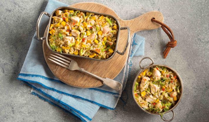 Foto em tons de cinza, azul e marrom uma bancada cinza vista de cima, um pano azul claro, uma tábua de madeira redonda com um recipiente retangular com alças que contém arroz com pedaços de frango, um garfo e um recipiente redondo com arroz e frango