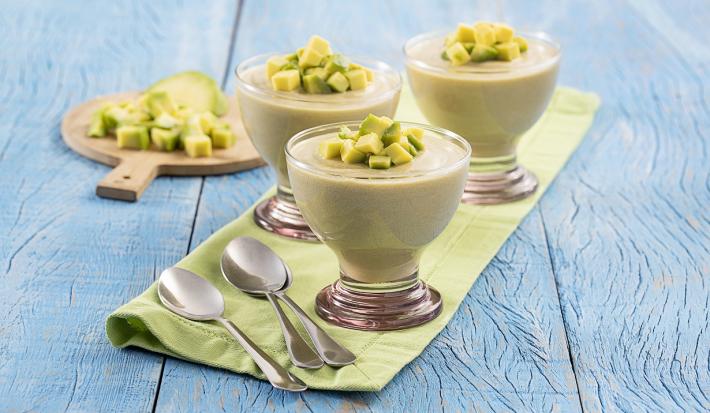 Foto da receita de mousse de abacate disposta em três taças de vidro com abacate em cubos para decorar, estão em cima de uma mesa azul com um pano verde separando, em cima do pano duas colheres prateadas. Ao fundo, abacate em cubos numa tábua de madeira