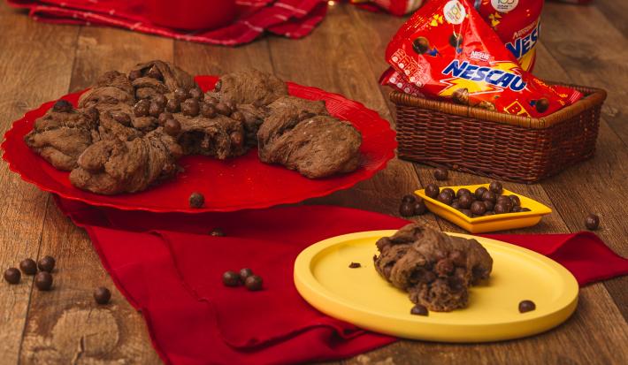 Foto de uma bancada de madeira. Sobre ela há um tecido vermelho, um prato amarelo com um pedaço de pão de Nescau, o pão maior ao fundo sobre um prato vermelho, uma cestinha com pacotes de Nescau Ball e algumas bolinhas de chocolate espalhadas pela mesa.