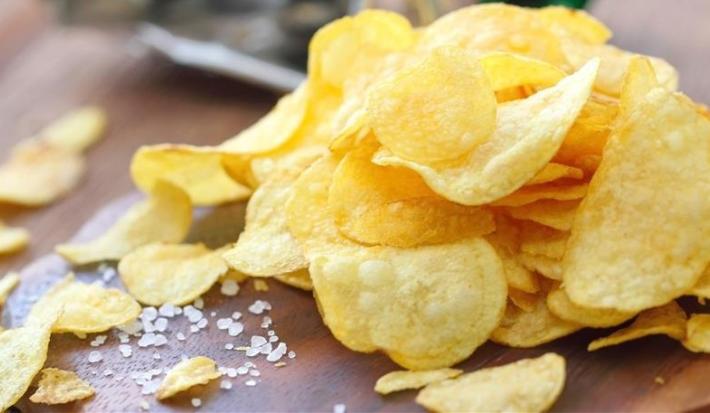 Fotografia de alguns chips de batatas sobrepostos sobre uma mesa de madeira escura, juntamente com um pouco de sal grosso. No fundo tem um saquinho verde.
