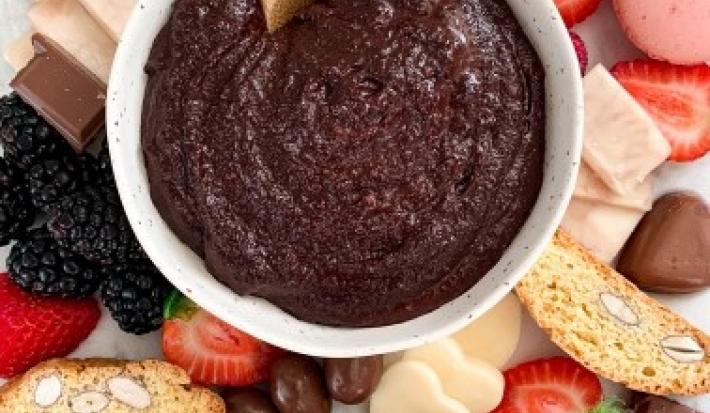 Fotografia em tons de vermelho e rosa em uma bancada com uma toalha colorida. Ao centro, um prato redondo fundo com o creme de chocolate de brownie com frutas, castanhas, frutas secas e pães ao redor dele.