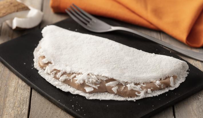 Fotografia em tons de preto, laranja e branco de uma bancada de madeira, sobre ela um prato retangular preto com a tapioca e um garfo. Ao fundo um paninho laranja com coco.