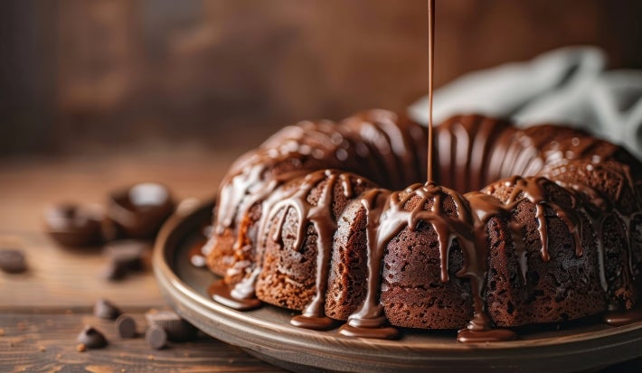 Fotografia de um bolo de chocolate em forma de bundt, coberto com chocolate derretido que escorre pelas laterais, posicionado em um prato escuro sobre uma superfície rústica.