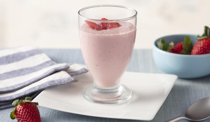 Fotografia em tons de azul e rosa de uma bancada com um prato quadrado, sobre ele um copo com o creme de morango e pedacinhos de morango. Ao redor um paninho listrado azul e branco e um potinho com morangos.