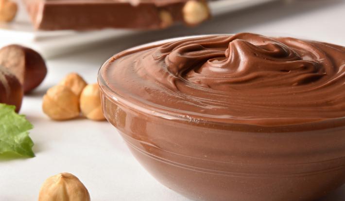 Fotografia em tons claros de um recipiente de vidro fundo com creme de avelã. O recipiente está sobre uma mesa branca com avelãs espalhadas.