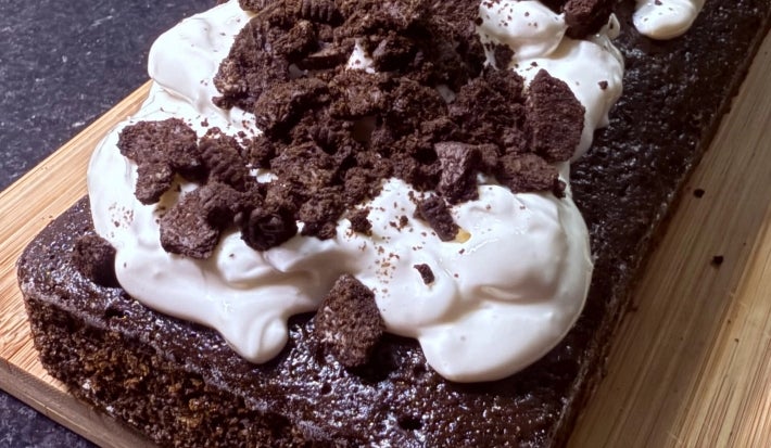 Fotografia de um bolo de negresco, assado na forma de bolo inglês, sobre uma tábua de madeira. O bolo está decorado com iogurte natural e biscoitos negresco quebrados.