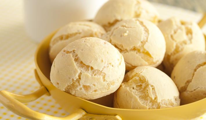 Fotografia em tons de amarelo em uma bancada de madeira esverdeada, um paninho branco com bolinhas amarelas, um recipiente amarelo com vários pães de queijo dentro dele. Ao fundo, uma xícara branca com café.