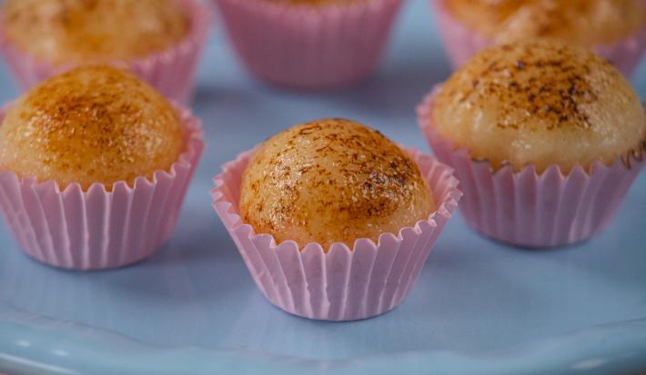 Fotografia de seis unidades de embalagens rosas, uma perto da outra, formando um hexágono. em cada forminha, tem um brigadeiro de creme brulée. As embalagens estão sobre um apoio de cor azul claro.