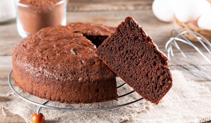 Imagem de um bolo redondo de chocolate com uma fatia em destaque. Compondo a imagem, um fuet, ovos e um pote com chocolate em pó.