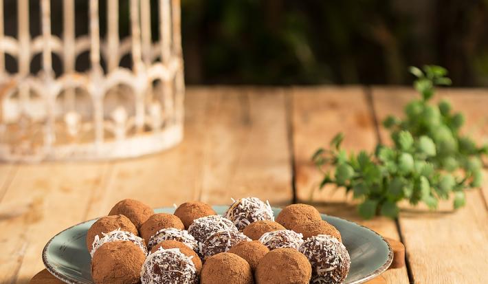Fotografia em tons de verde em uma bancada de madeira com um prato raso verde ao centro apoiado em uma tábua de madeira e os brigadeiros em cima. Ao lado, brigadeiros espalhados pela bancada.