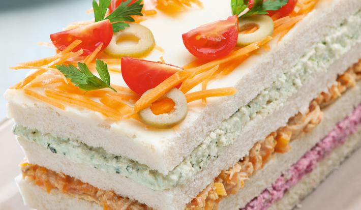 Fotografia do torta fria retangular, feita com pão de forma, e cores rosa, laranja e verdes no recheio, para decoração tomate cereja, cenoura e azeitona. A torta está sendo servida em um parto retangular rosa claro.