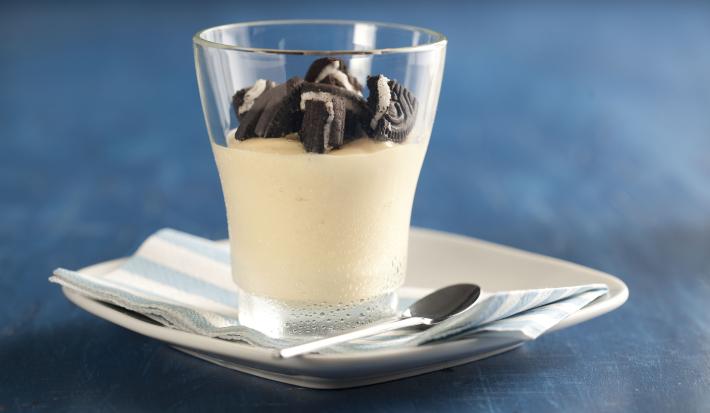 fotografia em tons de azul e branco de uma bancada azul vista de frente, contém um pratinho branco com um pano em tons de azul branco, um copo transparente com creme de maracujá com pedaços de biscoitos por cima.