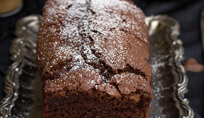 Fotografia em tons de marrom de uma bancada marrom vista de cima, contém uma bandeja de alumínio com um bolo inglês de chocolate com açúcar polvilhado por cima.
