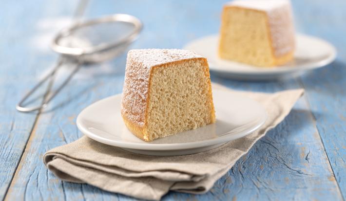 Foto da Receita de Mini Bolo de Baunilha. Observa-se duas fatias de bolo de baunilha, uma em cada prato branco sobre um guardanapo bege em uma superfície de madeira azul clara. Ao fundo, uma peneira com açúcar de confeiteiro de decoração.