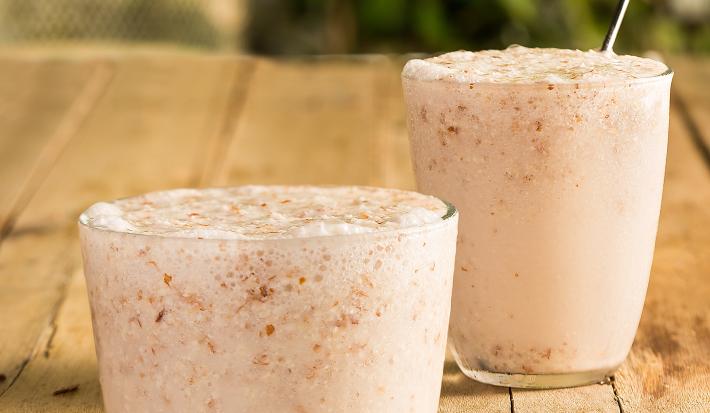 Fotografia em tons de marrom em uma mesa de madeira com dois copos de vidro com o frappé de amêndoas com coco dentro e um canudo de aço inox. Amêndoas espalhadas pela mesa.