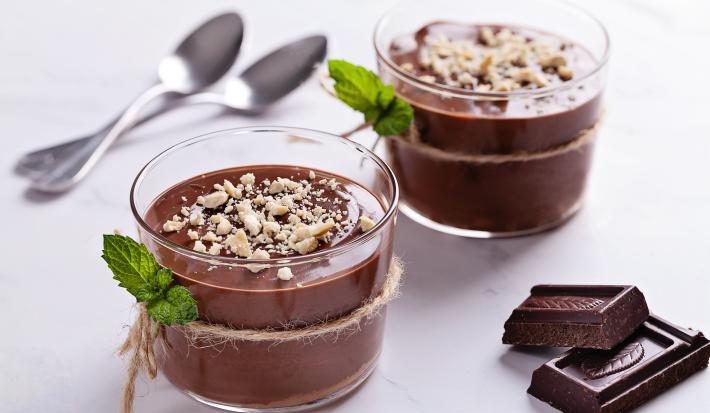 Fotografia de dois recipientes de vidro decorativos com pudim de café crocante com uma mini corda amarrando o pote e folhas de hortelã junto. Ao fundo, duas colheres de sobremesa, e ao lado, dois quadrados de chocolate amargo sobre uma mesa branca.