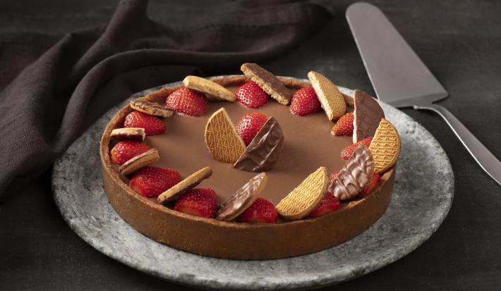 Fotografia em tons de preto, com fundo preto com paninho preto e prato cinza, com uma torta de chocolate, morangos e biscoitos Calipso e uma espátula prata.