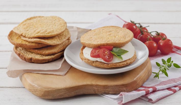 Fotografia em tons de branco, marrom e vermelho, ao centro fatias de pão sobre guardanapo e prato branco com lanche contendo queijo branco e tomate sobre tábua, ao lado guardanapo branco com listras vermelhas e tomates-cereja, tudo sobre bancada branca.