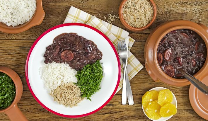 Fotografia em tons de marrom e vermelho de uma bancada de madeira, sobre ela recipientes redondos com feijoada, arroz, couve e farofa. Ao centro um paninho branco xadrez com um prato branco redondo com um prato feito de feijoada.