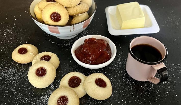 Foto da receita de mantecal com geleia em tons de vermelho servidos em um bowl de porcelana com adornos cor de rosa. À frente há alguns biscoitos mantecal soltos além de uma caneca de café e um recipiente com geleia e ao lado um recipiente com manteiga