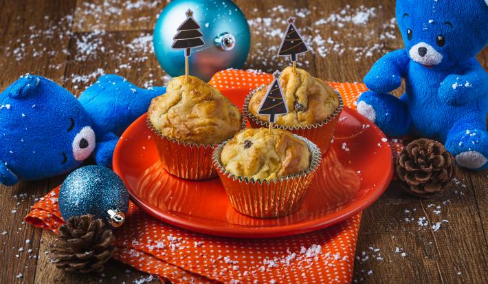 Fotografia em tons de laranja e azul em uma bancada de madeira, um prato laranja com três muffins em cima dele. Ao lado, enfeites e decoração natalina.