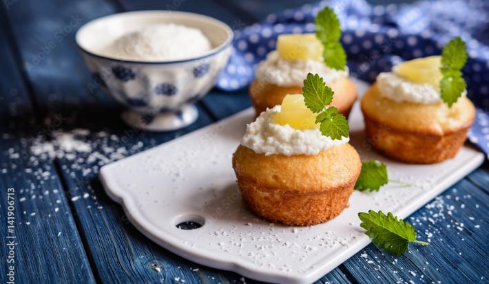 Fotografia de três muffins de abacaxi com decoração de um creme branco, abacaxi e folhas de hortelã sobre uma mini tábua na cor branca. Ao fundo, o creme branco dentro de um recipiente fundo nas cores azul e branco, sobre uma mesa de madeira na cor azul.