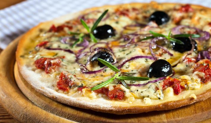 Fotografia de uma pizza de massa fina com mozarela, gorgonzola, requeijão, tomate seco, orégano, cebola roxa e azeitonas pretas. A pizza está sobre um apoio de madeira.
