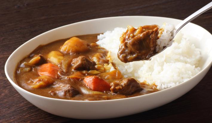 Fotografia de um recipiente branco fundo oval sobre uma mesa de madeira escura. Dentro do recipiente tem uma receita de kare de carne com legumes e arroz branco. Sobre o recipiente tem uma colher de sopa segurando um pouco da receita.