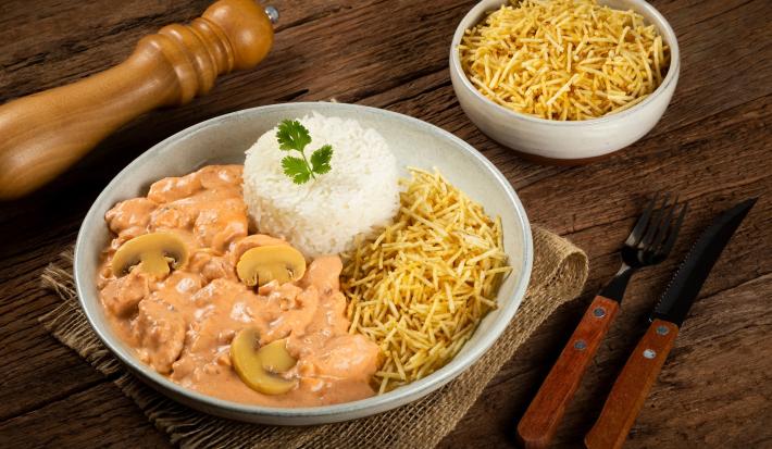 Imagem de um prato com arroz, batata palha e strogonoff de frango. Ao lado, um pote de batata palha e um pimenteiro.