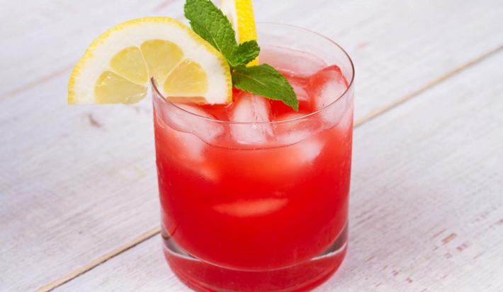 Fotografia em tons de vermelho em uma bancada de madeira de cor branca. Ao centro, um copo contendo a bebida pink lemonade.