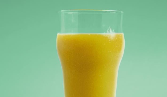 Fotografia em tons de verde em um fundo e bancada verde com um copo de vidro e o suco de manga, limão-siciliano e erva-cidreira dentro dele.