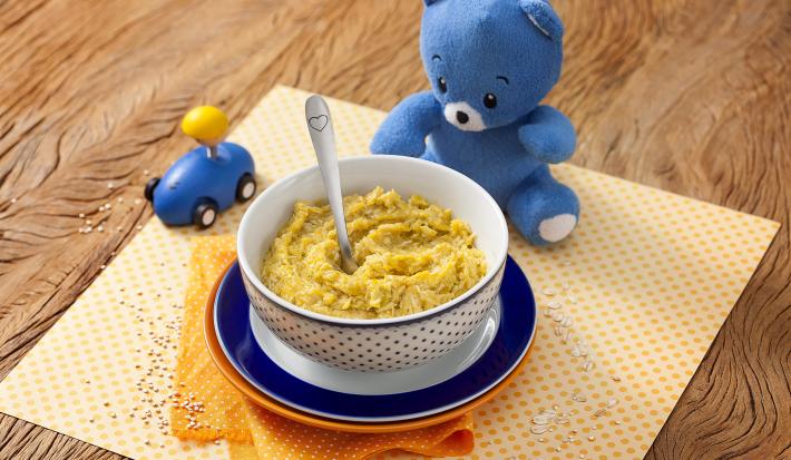Fotografia em tons de laranja em uma bancada de madeira com um paninho amarelo de bolinha no centro e um potinho branco com bolinhas azuis com a sopinha de frango com abóbora. Ao lado, um ursinho de pelúcia azul e um carrinho azul de brinquedo.