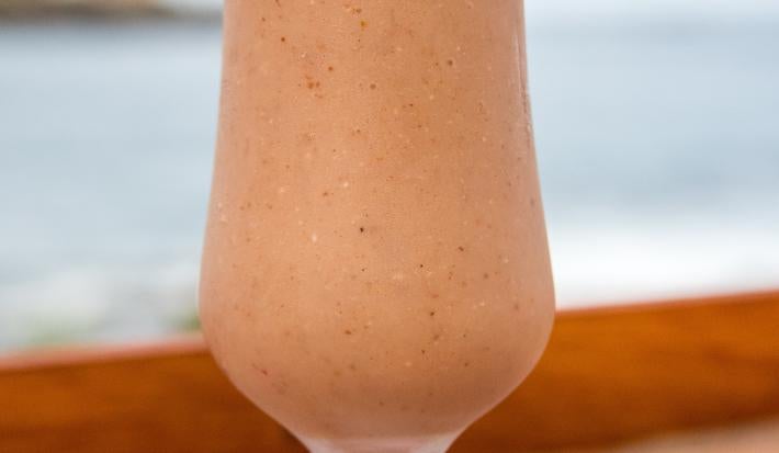 Fotografia de um copo de vidro, estilo taça, com uma bebida de suco de goiaba e neston sobre uma mesa de madeira com vista para a praia.