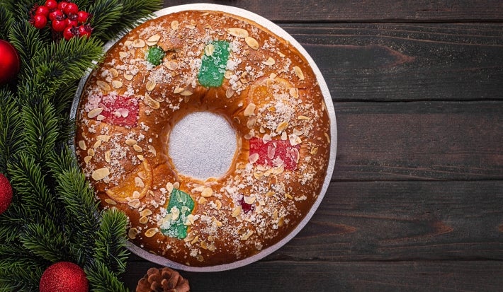 Fotografia de um bolo de reis enfeitado com frutas cristalizadas em vermelho, verde e laranja, polvilhado com açúcar e amêndoas. Colocado em um prato metálico, com ramos de pinheiro e enfeites vermelhos ao fundo.