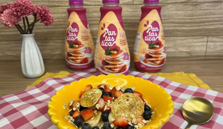 Fotografia em tons de rosa e amarelo com um prato amarelo ao centro. Dentro do prato existe um cereal feito de mini panquecas, frutas vermelhas, aveia e mel. Ao fundo existe três garrafas de Pantásticas Zero Açúcares.
