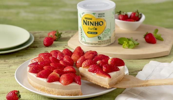 Fotografia em tons de verde e vermelho de uma bancada, sobre ela um prato redondo com uma sobremesa com base de biscoito, recheio de creme branco e morangos decorando. Ao fundo uma lata de NINHO Origem Vegetal e morangos.