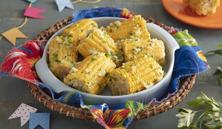 Foto da receita de Espiga de Milho Temperada. Observa-se um recipiente de porcelana branco com 7 espigas de milho cortadas ao meio e temperadas dentro. A foto está decorada com bandeirinhas de festa junina e tecidos temáticos